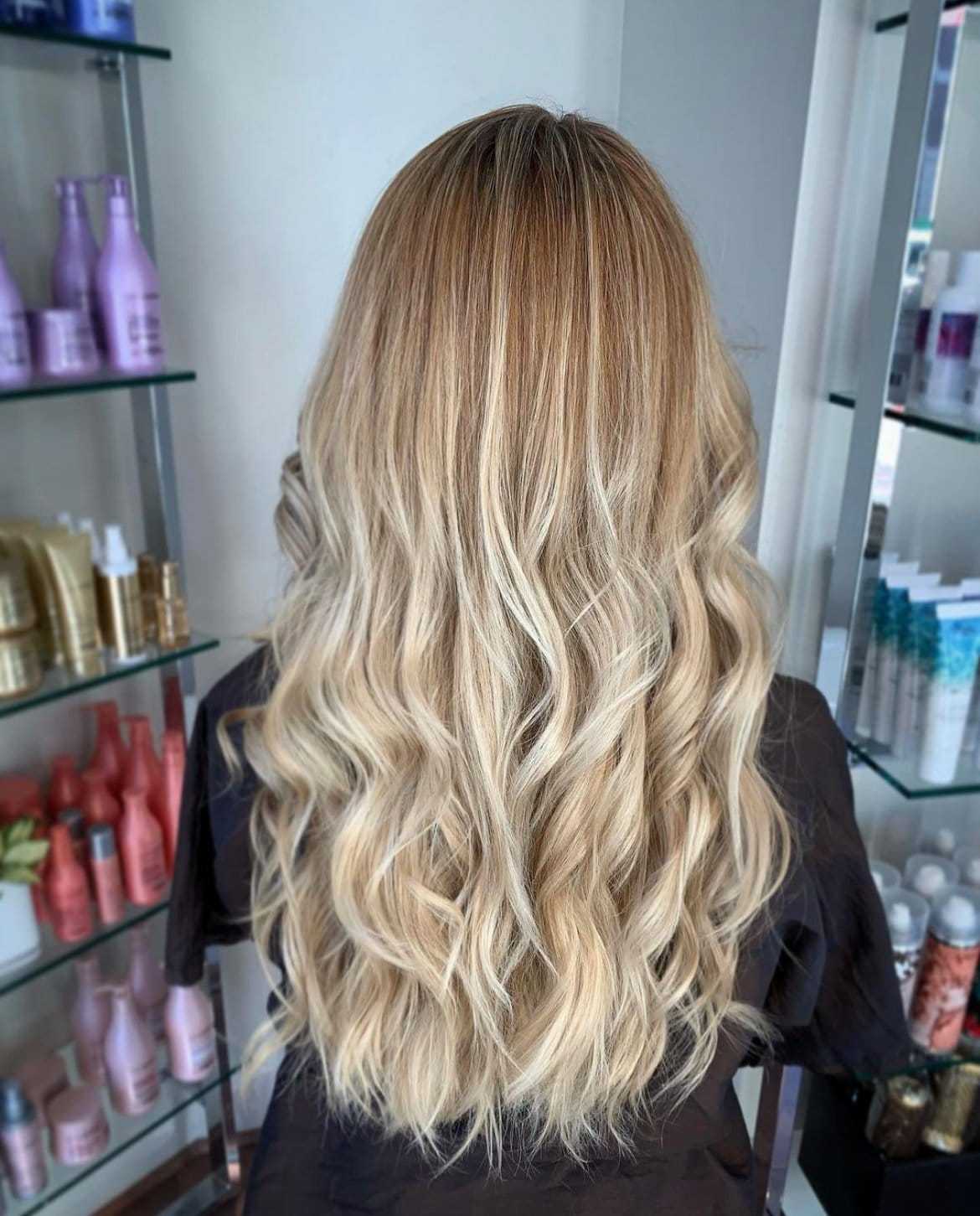 Wavy blonde hair styled in soft curls, displayed against a salon backdrop with hair products.